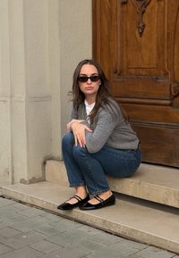 Γκρίζα καζάκα πάνω από λευκό πουκάμισο, μπλε τζιν και μαύρες παναδόλες από πατενταρισμένο δέρμα με σταυρωμένα λουριά, καθισμένη σε μια πέτρινη σκάλα κοντά σε μια ξύλινη πόρτα.