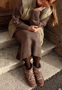 Vestido de punto marrón con dobladillo acanalado, combinado con un chaleco acolchado verde claro y botas marrones hasta el tobillo con acentos texturizados.