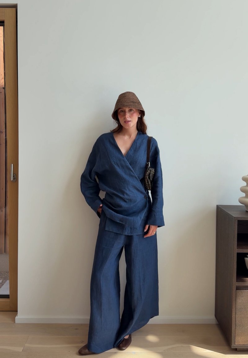 Femme portant un top cache-cœur bleu et un pantalon ample, des chaussures marron et un chapeau marron, debout contre un mur blanc, la main dans la poche.