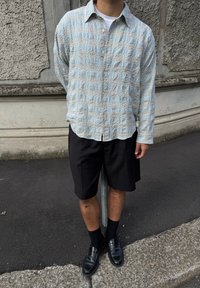 Šviesiai mėlynas ir smėlio spalvos raštuotas marškinėlių mygtukų užsegimas su laisvu siluetu, derinamas su juodomis prisiūtomis šortais ir blizgiais juodais loaferiais.