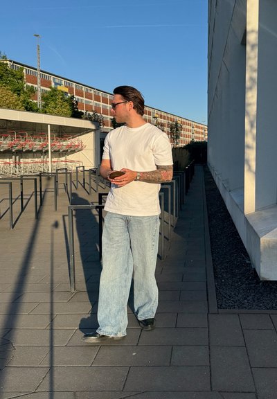 Camiseta blanca, jeans de pierna ancha azul claro y zapatos negros. De pie al aire libre con carritos de compra y un edificio moderno al fondo.