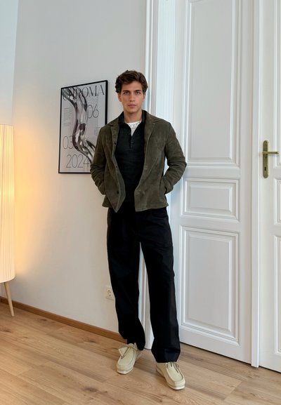 Chaqueta de ante verde oliva, suéter oscuro, pantalones negros y calzado beige claro. De pie en una habitación luminosa con una puerta y obras de arte.
