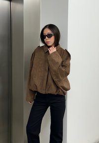 Veste oversized en suédine marron avec col et fermeture éclair avant, associée à un pantalon noir. Le mannequin porte des lunettes de soleil noires et se tient près d'un ascenseur.