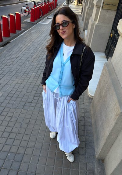 Mujer con el pelo largo que lleva gafas de sol, un cárdigan azul, chaqueta negra, falda blanca y zapatillas deportivas, de pie en una acera de la ciudad cerca de puestos de alquiler de bicicletas.