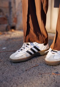 Bianche sneakers con strisce nere, dettagli in pelle scamosciata e una suola in gomma grigia testurizzata. Indossate con pantaloni marroni leggermente larghi su ghiaia.
