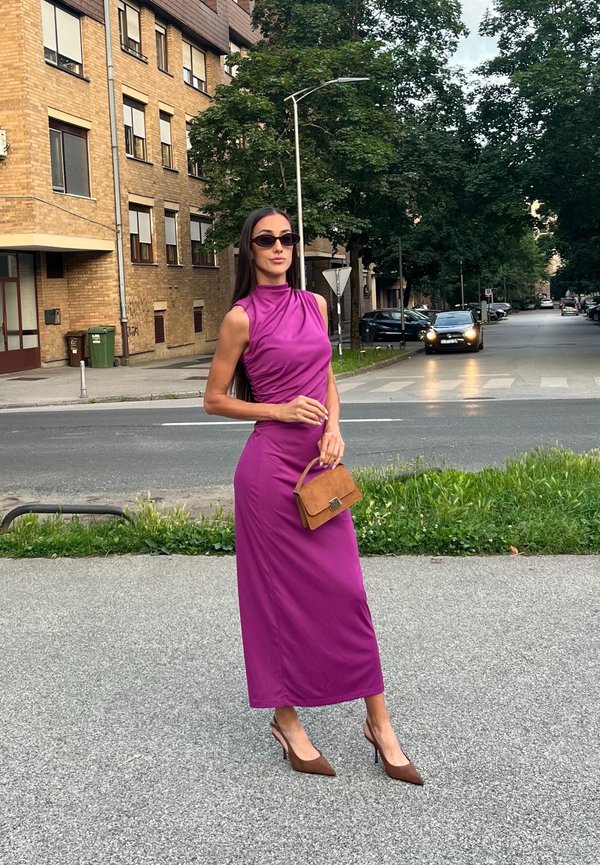 Femme en robe violette sans manches et talons marron tenant un sac à main marron, debout sur un trottoir en ville avec des arbres et des bâtiments en arrière-plan.