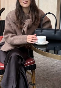 Hnedý vlnený sveter s detailom uzla, čierna kabelka a biely pohár na kávu na sklenenom stole, sediac na prútenej stoličke s červenými akcentmi.