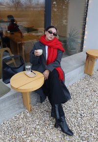 Un manteau gris est porté sur une chemise blanche, assorti d'une écharpe rouge vif et de bottes à talons noires. Une table en bois porte une tasse et un verre d'eau.