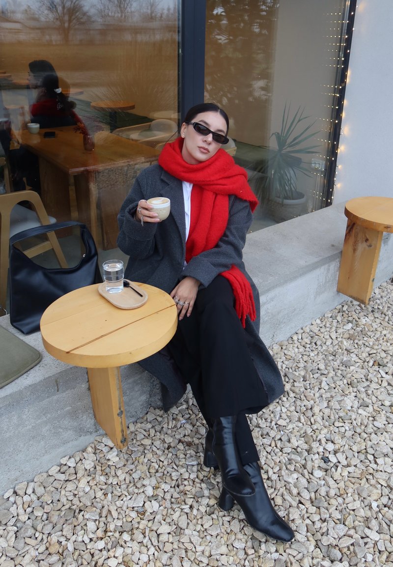 Un manteau gris est porté sur une chemise blanche, assorti d'une écharpe rouge vif et de bottes à talons noires. Une table en bois porte une tasse et un verre d'eau.