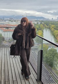 Brauner Pelzmantel mit weitem Kragen, kombiniert mit braunen Schlaghosen und dunklen Sneakers. Hält eine dunkle Tote-Tasche. Holzboden und Stadtansicht im Hintergrund.