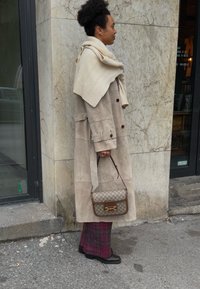Manteau trench en daim beige avec une écharpe en tricot crème drapée sur les épaules. Sac à main marron avec détail de logo, pantalon à carreaux et chaussures noires.