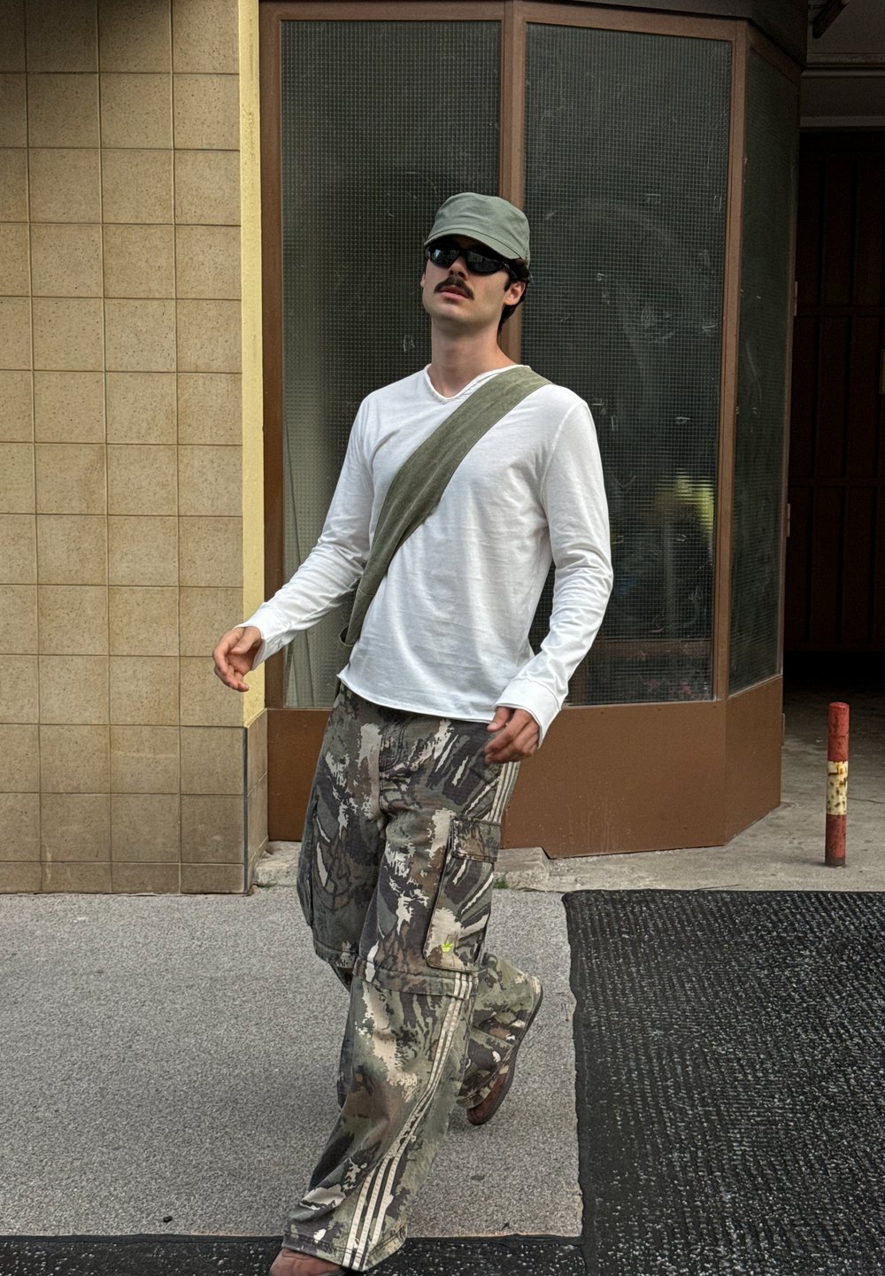 Weisses Langarmhemd, olivgrüne Umhängetasche, gemusterte Cargo-Hosen in Beigetönen und Oliv, braune Sandalen, Sonnenbrille, Baseballkappe.