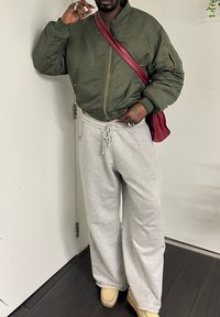 Personne portant une veste bomber vert olive, un pantalon de survêtement gris, des chaussures beige, et portant un sac bandoulière bordeaux, debout à l'intérieur près d'une porte blanche.