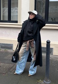 Schwarzer langer Mantel über einem schwarzen Hoodie, beigefarbenes kariertes Hemd in der Taille gebunden, hellblaue Jeans mit Used-Effekt und beigefarbene spitze High Heels. Schwarze Handtasche.
