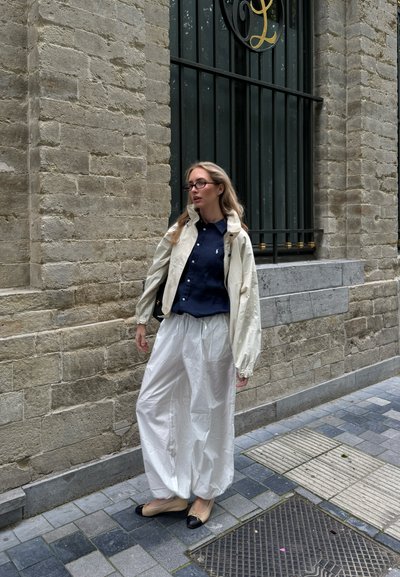 Joven mujer con gafas que lleva una camisa azul marino, chaqueta crema y pantalones blancos y sueltos, de pie en una calle adoquinada junto a una pared de ladrillos.
