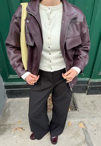 Bordeauxrode leren jas met zilveren rits, crèmekleurige gebreide trui met knopen, wijde zwarte broek en bordeauxrode schoenen. Gele tas als accent.