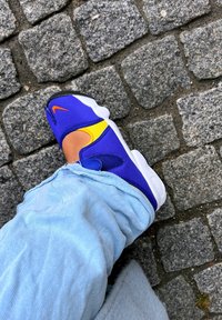 Stopalo u plavim i žutim Nike čarapastim cipelama s narančastim swooshom, svijetloplavim trapericama, na sivom kameniku.