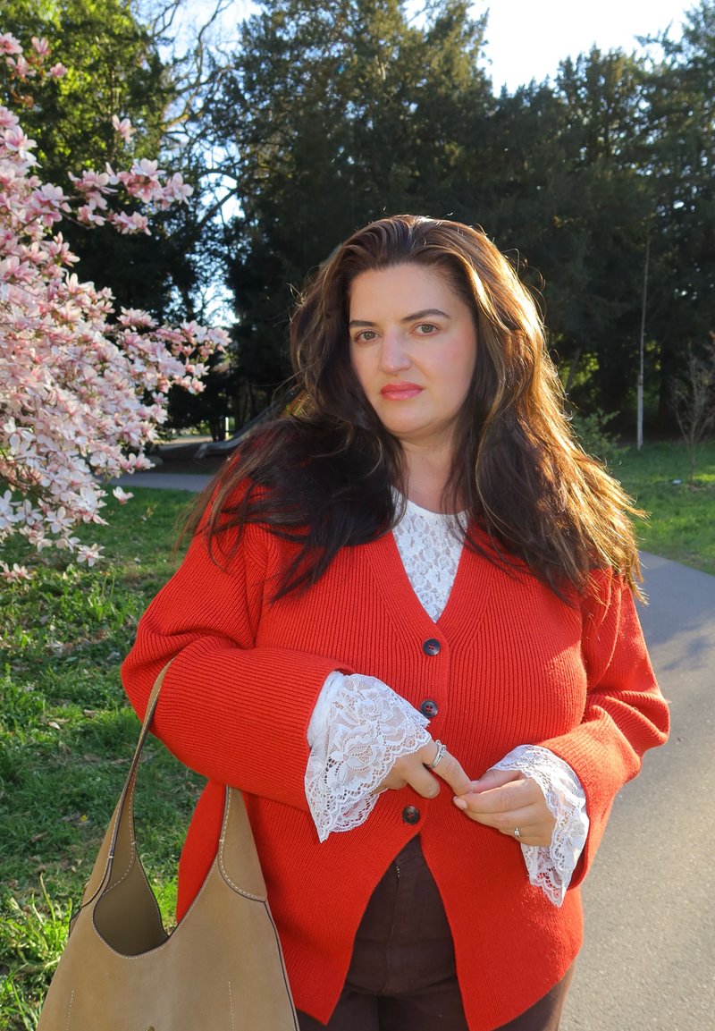 Femme aux longs cheveux bruns portant un cardigan orange avec des manches en dentelle blanche, debout près de fleurs roses et tenant un sac beige en plein air.