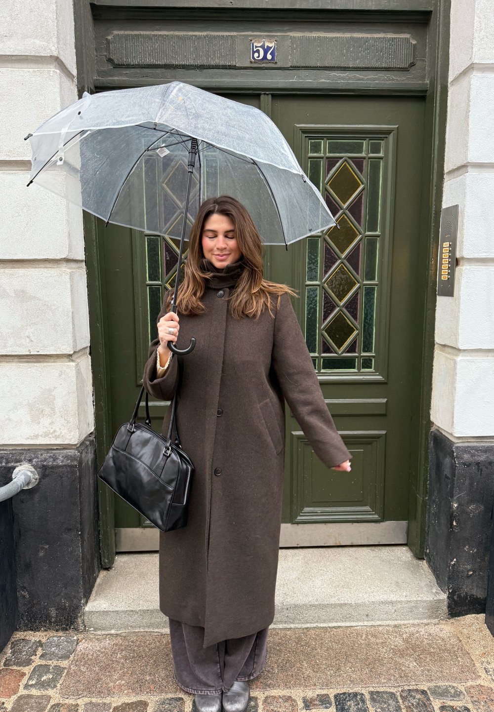 Manteau en laine marron avec de gros boutons, pantalon gris à jambes larges, sac à main noir, et parapluie transparent devant une porte verte avec des accents en verre.