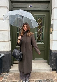 Manteau en laine marron avec de gros boutons, pantalon gris à jambes larges, sac à main noir, et parapluie transparent devant une porte verte avec des accents en verre.