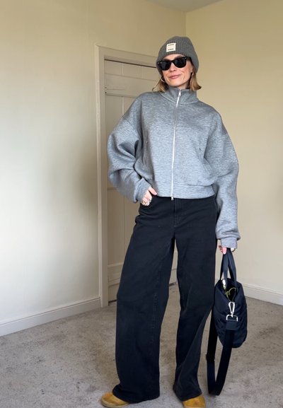 Persona con gafas de sol, gorro gris, sudadera gris con cremallera, pantalones negros anchos, sosteniendo una bolsa negra en una habitación interior sencilla.