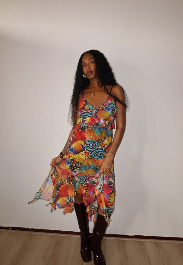 Maxikleid mit lebhaften Blumen- und Schmetterlingsmustern in mehreren Farben. Leichter Stoff, gerüschter Saum, Trägerdesign, kombiniert mit kniehohen braunen Stiefeln.