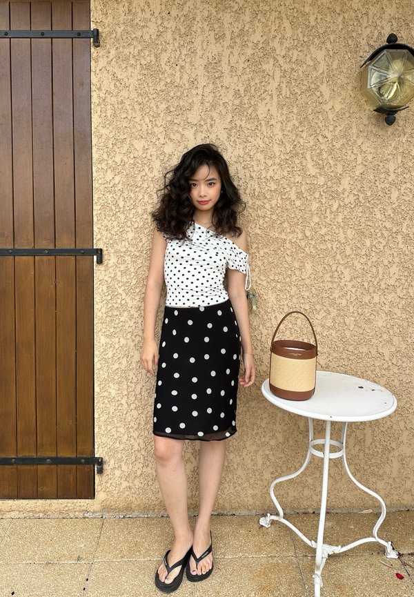 Jeune femme aux cheveux bouclés porte un top blanc à une épaule et une jupe noire à pois, debout à côté d'une petite table blanche avec un sac à main.