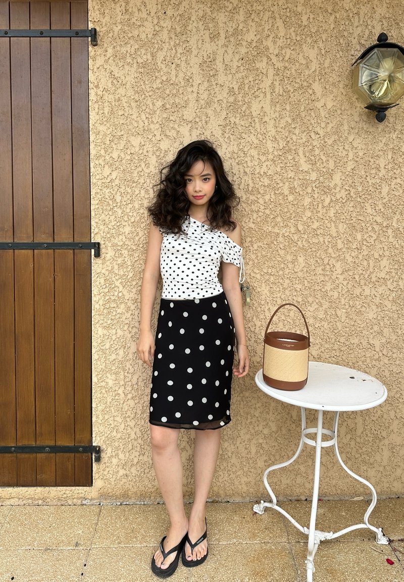 Young woman with curly hair wears a white one-shoulder top and black skirt with polka dots, standing next to a small white table with a handbag.