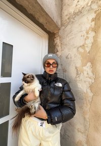 Μαύρο μπουφάν με λογότυπο, ανοιχτό γκρι σκουφάκι, πορτοκαλί γυαλιά ηλίου, κρέμα παντελόνι φόρμας. Κρατάει μια αφράτη λευκή γάτα με καφέ επισημάνσεις.