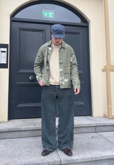 Hombre con gorra azul, chaqueta de camuflaje verde, suéter beige, jeans oscuros anchos y zapatos marrones, de pie en unos escalones de piedra frente a una puerta oscura.