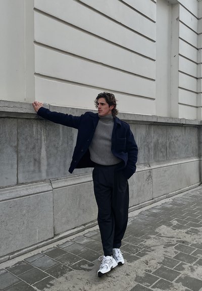 Abrigo azul marino sobre un suéter de cuello alto a rayas grises, combinado con pantalones negros y zapatillas blancas. De pie contra una pared clara.