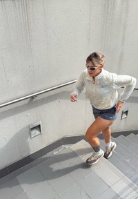 Moteris saulės akiniams, vilkinti baltą striukę ir šortus, bėga kylanti plytelių laiptais su metaliniu turėklu prie betono sienos.