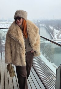 Femme en manteau de fourrure beige et chapeau avec des lunettes de soleil, debout sur un balcon enneigé avec vue sur une rivière et le paysage urbain.