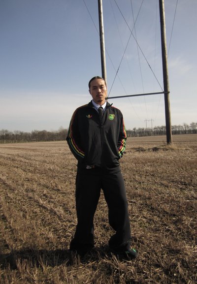 Hombre con chaqueta negra de Adidas con rayas verde, roja y amarilla, de pie en un campo cosechado cerca de altos postes de electricidad bajo un cielo despejado.