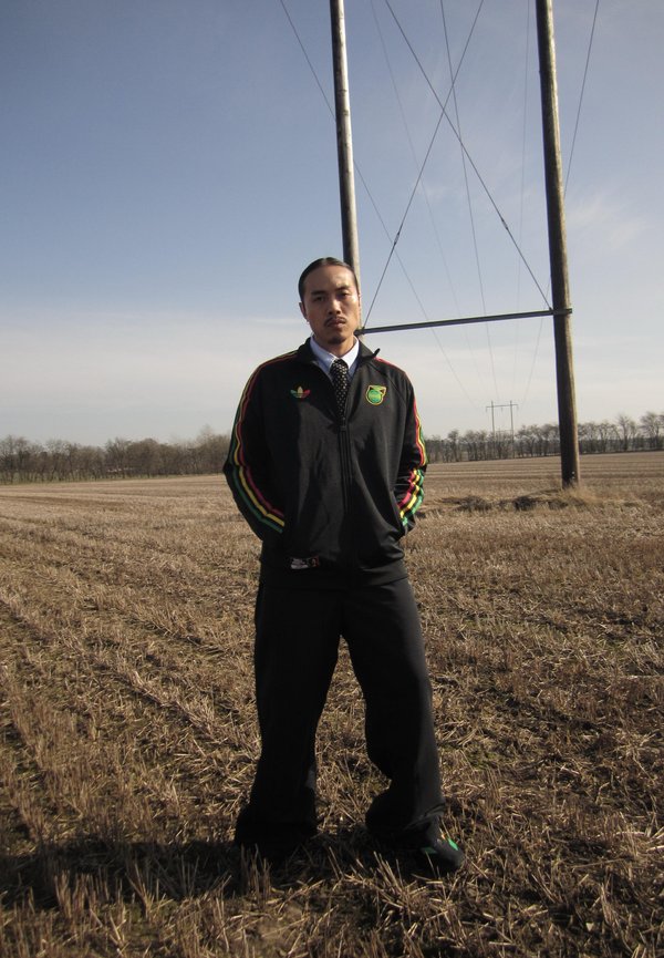 Mann trägt schwarze Adidas-Jacke mit grünen, roten und gelben Streifen und steht in einem abgeernteten Feld in der Nähe hoher Strommasten unter klarem Himmel.