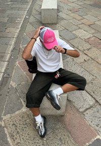 Gorra de malla rosa con texto, camiseta blanca de manga corta, pantalones cropped negros, calcetines blancos, zapatillas gris y negras, llavero con acentos de colores.