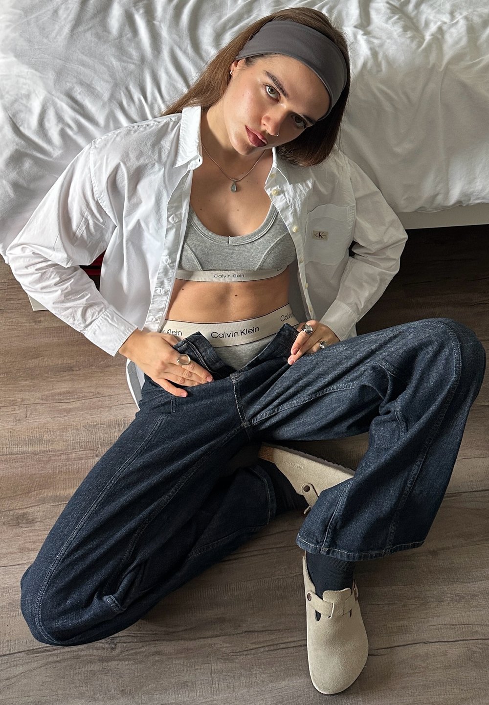 Hvit skjorte med knapper, grå sports-BH og undertøy, mørk denim med vide ben, beige tøfler, lagdelte ringer, lyst brunt hår, nøytral bakgrunn.