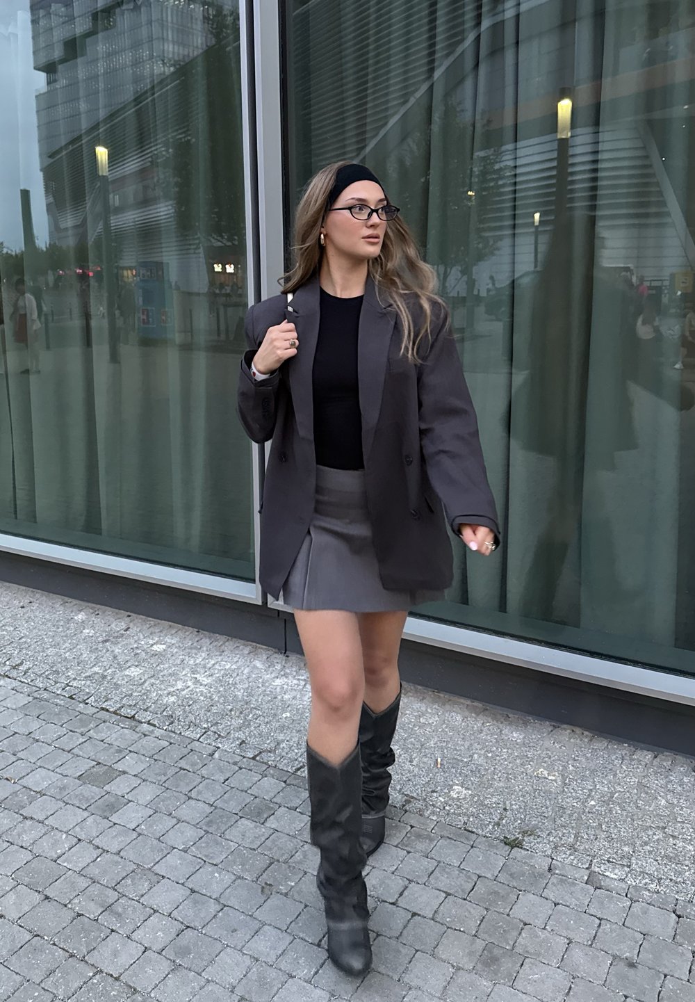 Szürke, oversized blézer egy fekete felsőn, párosítva egy szürke miniszoknyával és térdig érő fekete csizmával. Az öltözet letisztult dizájnnal és textúrákkal rendelkezik.