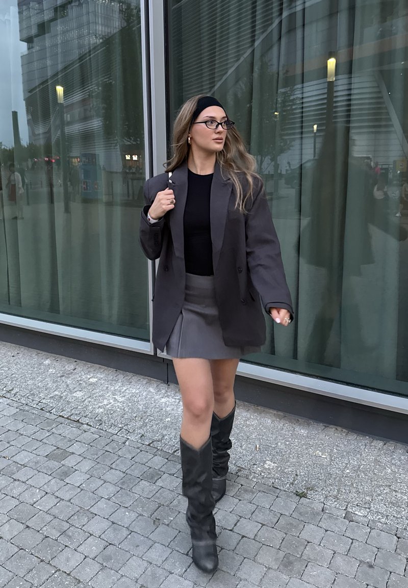 Grigia giacca oversize sopra una maglia nera, abbinata con una minigonna grigia e stivali neri fino al ginocchio. L'outfit presenta un design elegante e texture raffinate.