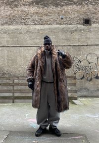Homme portant un manteau en fausse fourrure surdimensionné, un survêtement gris, un bonnet noir, des gants et des lunettes de soleil, debout sur un trottoir urbain avec un mur de graffiti en arrière-plan.