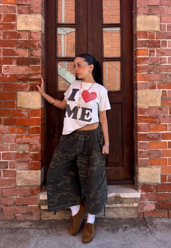 Jeune femme avec des lunettes se tient contre une porte en bois dans un mur de briques, portant un t-shirt blanc « I love me » et un pantalon large camouflage.