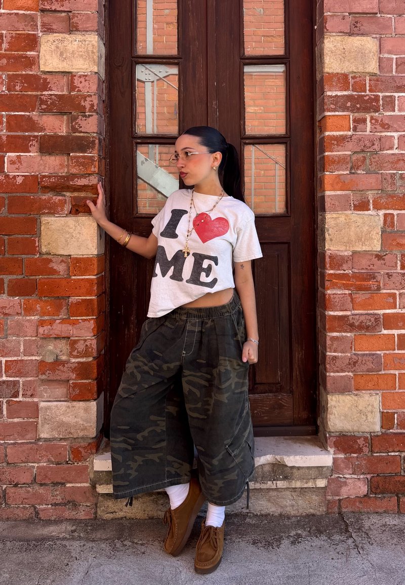 Jauna sieviete ar brillēm stāv pie koka durvīm ķieģeļu sienā, valkājot baltu T-kreklu ar uzrakstu „I love me“ un plašas maskēšanās krāsas bikses.