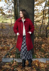 Palton roșu din lână cu nasturi negri, purtat peste un pulover tricotat crem și o fustă cu carouri alb-negru, completat de cizme negre până la gleznă.