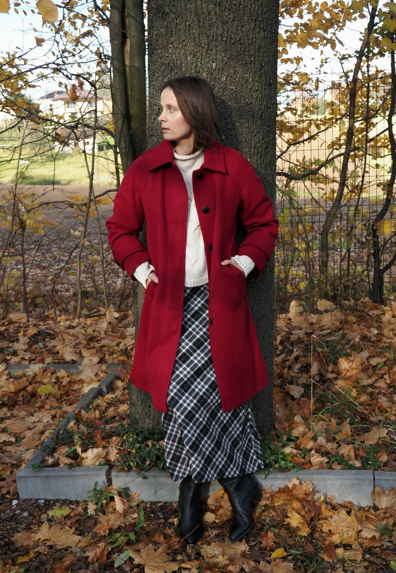 Roter Wollmantel mit schwarzen Knöpfen, getragen über einem cremefarbenen Strickpullover und einem schwarz-weißen karierten Rock, ergänzt durch schwarze Ankle Boots.