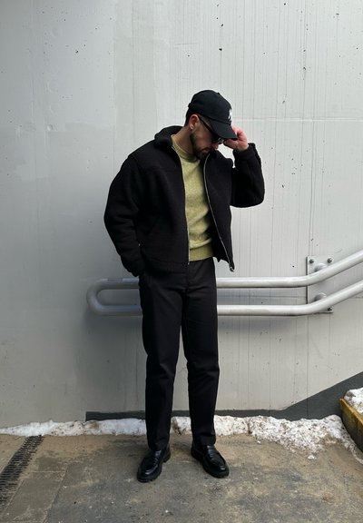 Chaqueta de forro polar negra con cierre en la parte delantera, suéter verde debajo, pantalones negros y zapatos negros. De pie contra una pared gris con nieve.