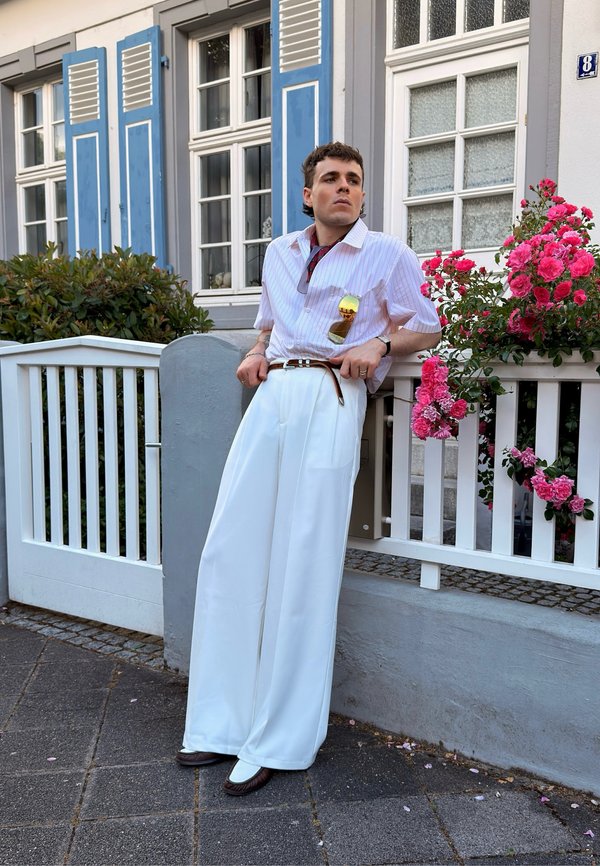 Personne portant un pantalon blanc à jambes larges et une chemise rayée, appuyée sur une clôture blanche à côté de fleurs roses en fleurs à l'extérieur d'une maison avec des volets bleus.