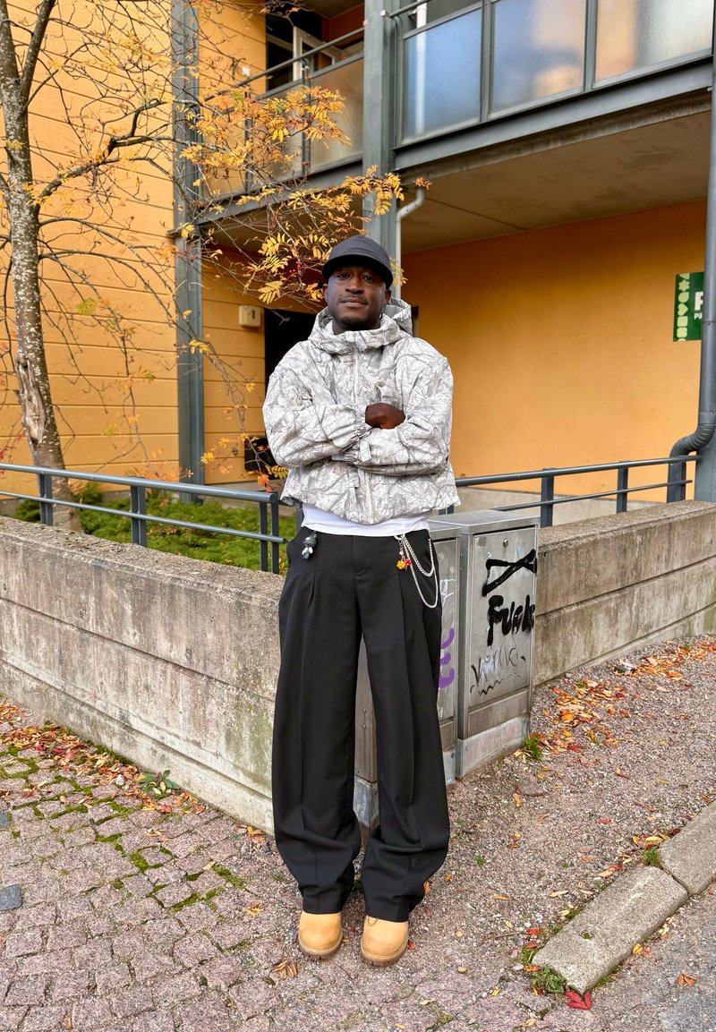 Veste camouflage grise, pantalon noir ample et bottes marron clair. Debout contre un mur en béton, entouré de feuilles d'automne, cadre urbain.