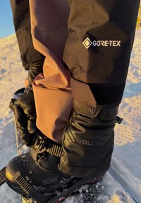 Črna Gore-Tex jakna s rožnato notranjostjo, v kombinaciji z teksturiranimi črnimi rokavicami z blagovno znamko Volcom, nad črnimi smučarskimi čevlji.