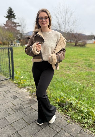 Chaqueta de piel marrón con forro beige, suéter beige claro, pantalones acampanados negros y zapatillas blancas en una zona pavimentada con césped.
