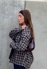 Bordó lakkbőr kézitáska ívelt formával és aranyszínű fém díszítésekkel, kockás inggel viselve. A háttérben szürke betonfal látható.